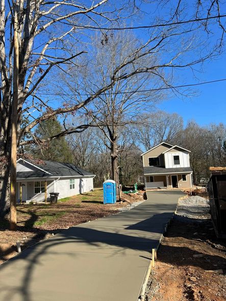 New concrete driveway leads to two houses under construction, blue sky, trees and portable toilet present.