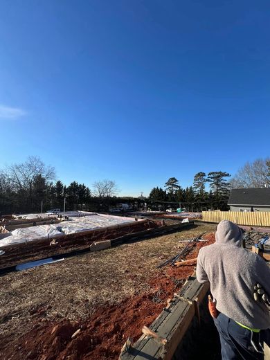 Construction site under a clear blue sky; worker in hoodie, dirt, wood, snow, trees in background.