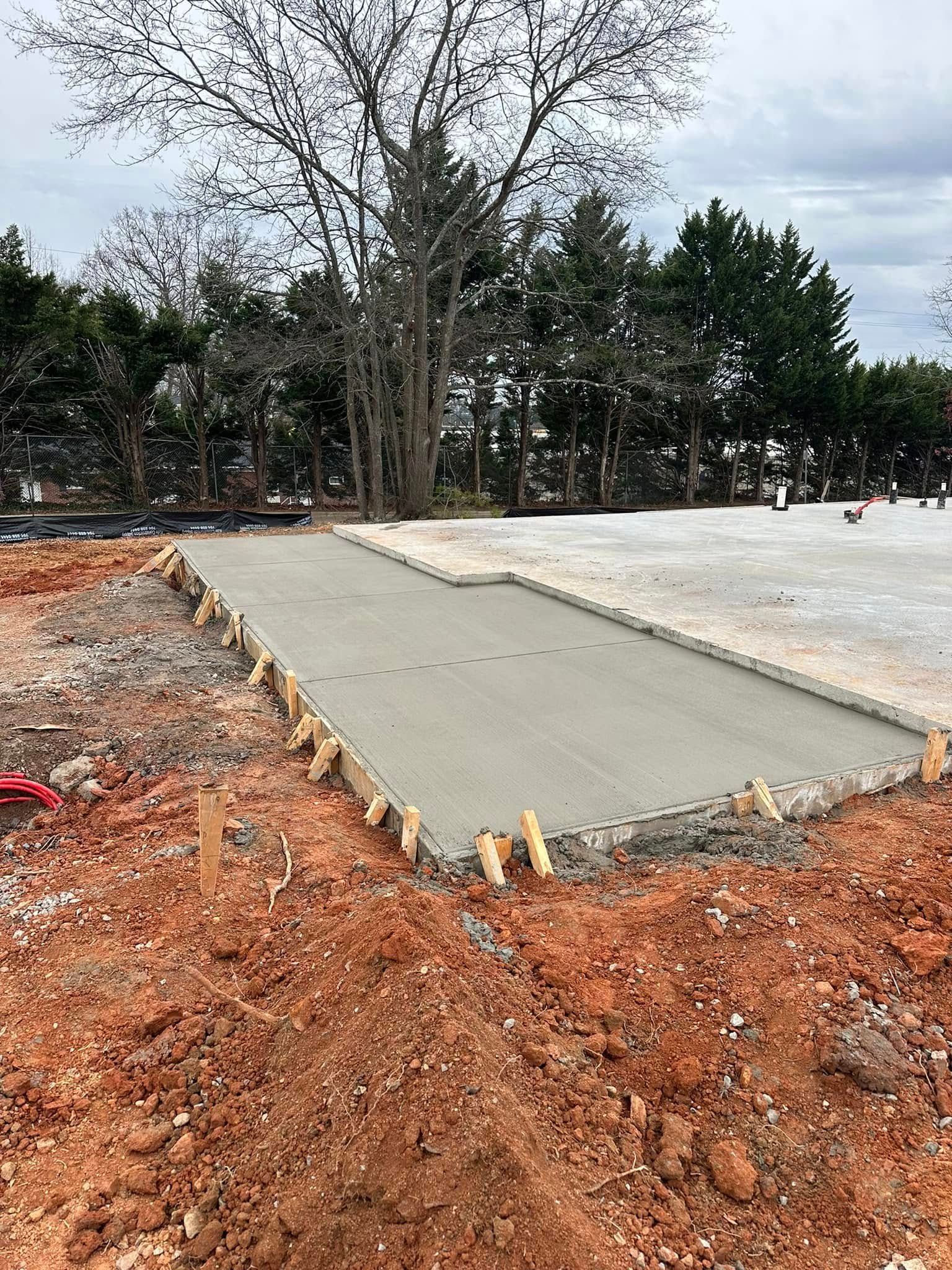 Construction site with a freshly poured concrete slab framed by wooden stakes, surrounded by red soil.