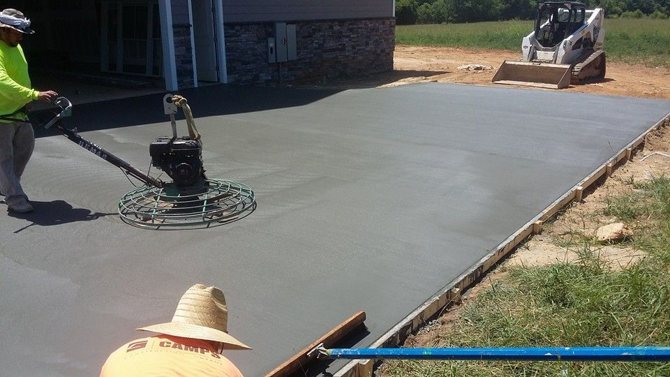 Workers smoothing fresh concrete driveway with a power trowel near a building and a small bobcat on a sunny day.