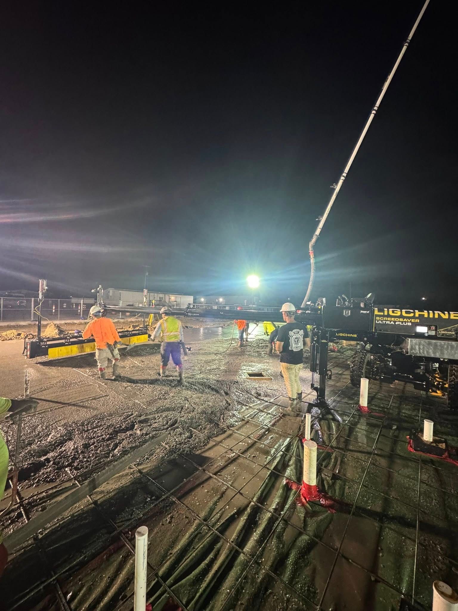 Construction workers pouring concrete at night. Bright floodlights illuminate the work site.