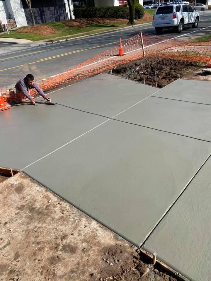 A worker smoothing wet concrete sidewalk sections with a trowel, near a road.