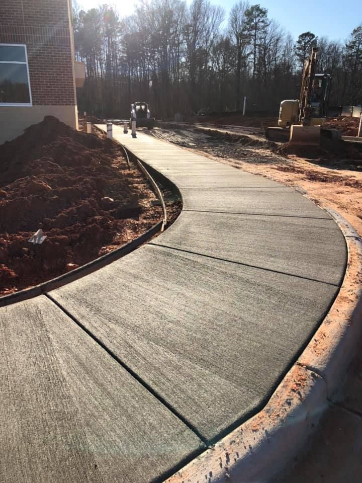 Curved concrete sidewalk under construction; brown soil, trees, and construction equipment visible.
