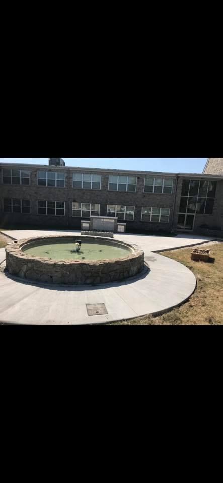 Stone fountain in a circular concrete patio, with a two-story brick building in the background.