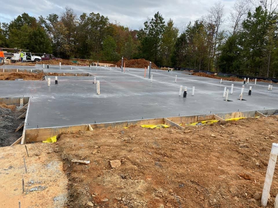 Newly poured concrete foundation with plumbing, surrounded by dirt, under a cloudy sky.