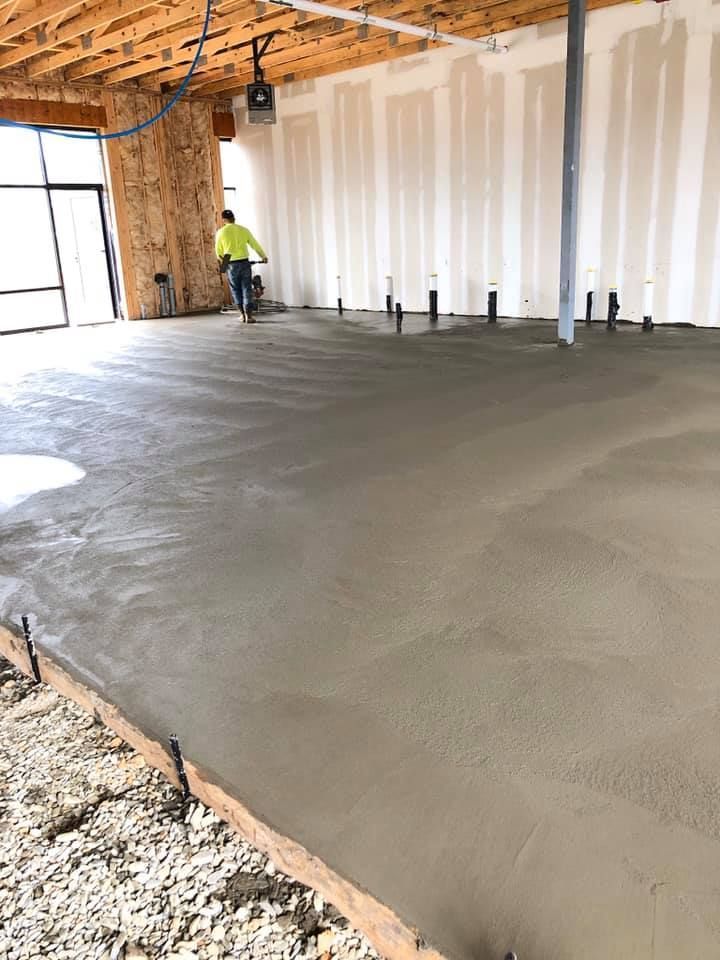 A worker spreads wet concrete on a construction site floor. The interior space has exposed beams and unfinished walls.