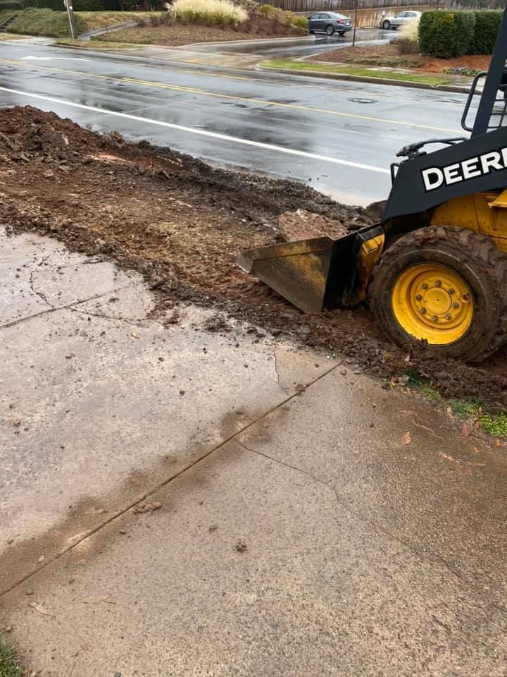 A small John Deere skid steer loader pushing dirt onto a sidewalk next to a wet street.