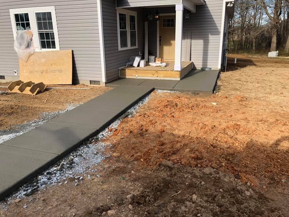 Newly poured concrete ramp leading to a house entrance. House is light gray, surrounded by dirt.