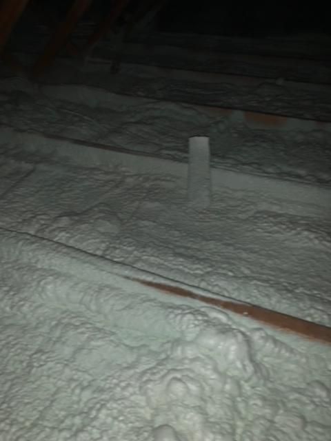 Une isolation en mousse pulvérisée blanche remplit un grenier, recouvrant les tuyaux et les poutres en bois.