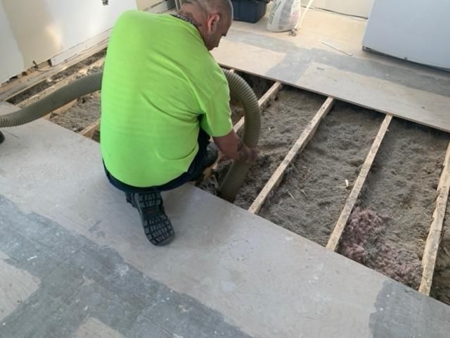 Un homme en chemise verte agenouillé utilise un tuyau pour remplir une cavité de plancher avec de l'isolant, à l'intérieur.