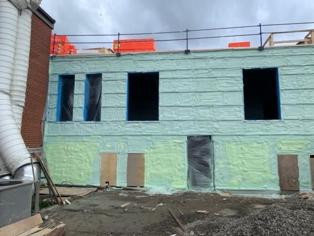 Extérieur du bâtiment en construction avec isolation verte, fenêtres ouvertes et porte partiellement installée.