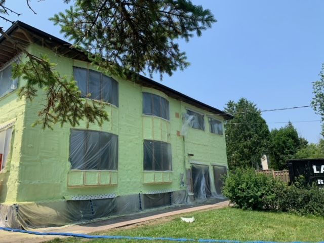 Bâtiment de deux étages avec isolation verte en cours de travaux, recouvert de bâche en plastique, journée ensoleillée.