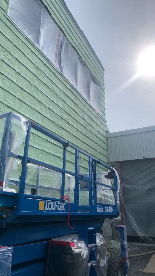 Un élévateur à ciseaux bleu à côté d'un bâtiment avec un revêtement vert, des fenêtres recouvertes d'une pellicule plastique, une journée ensoleillée.