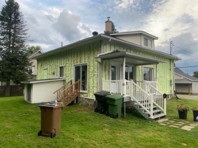 Vue latérale d'une maison à deux étages avec un revêtement vert clair, des escaliers de porche blancs et des poubelles vertes sur la pelouse.