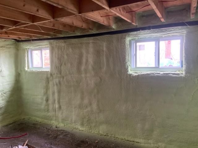 Mur intérieur du sous-sol avec isolation en mousse pulvérisée autour de deux fenêtres, avec poutres en bois au-dessus.