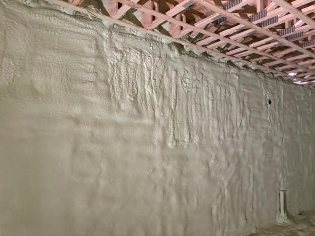 Isolation en mousse pulvérisée sur un mur de sous-sol, sous des poutres de plafond en bois apparentes ; une couleur beige clair.