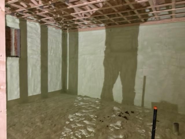 Intérieur d'une pièce avec isolation en mousse projetée sur les murs. On distingue les ombres d'une personne et des poutres. Le sol est irrégulier.