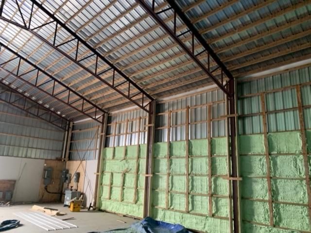 Intérieur d'un bâtiment en construction, avec isolation en mousse pulvérisée verte sur les murs, fermes métalliques au-dessus et toiture métallique.