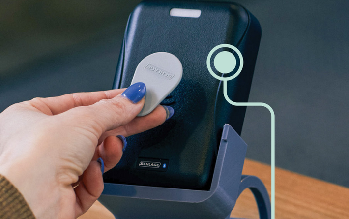 A hand holding a gray key fob to a digital terminal on a stand, with a circular icon indicating a contactless scan.