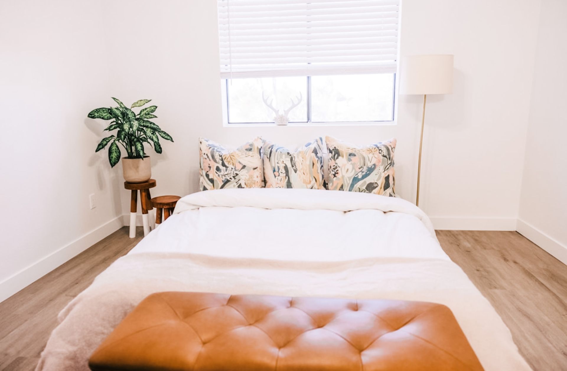 Bedroom with a bed, ottoman, plant, window, and floor lamp. White walls and a neutral color palette.