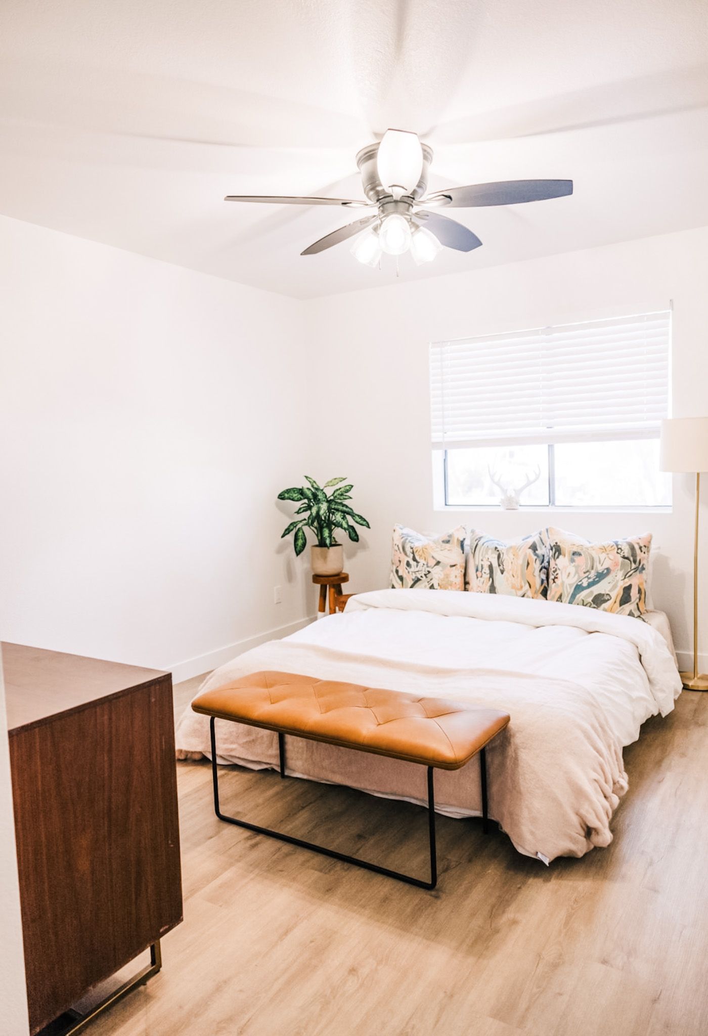 Bedroom with a bed, bench, wooden dresser, and a plant in front of a window.