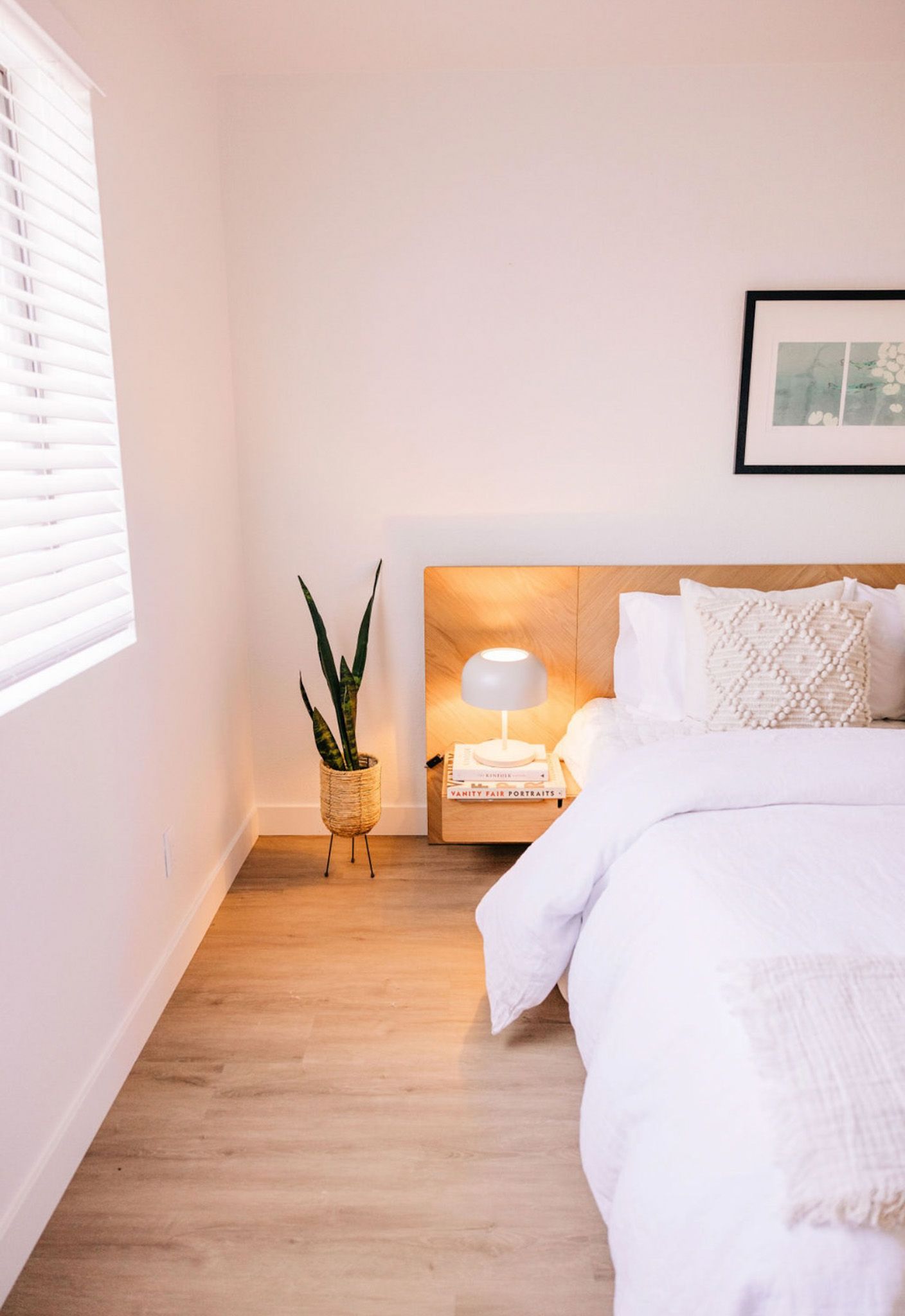 Bedroom with a bed, nightstand, plant, window, and artwork. Neutral tones with natural light.