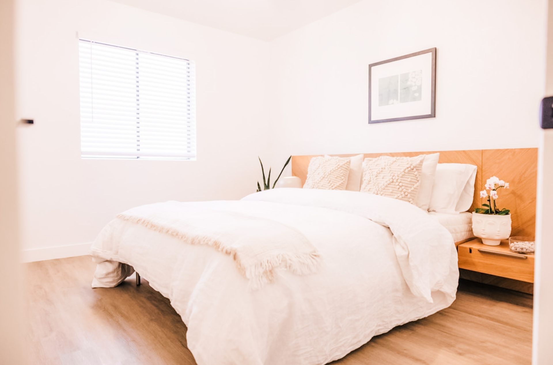 Bright bedroom with a bed, nightstand, window, and artwork. Mostly white with wooden accents.