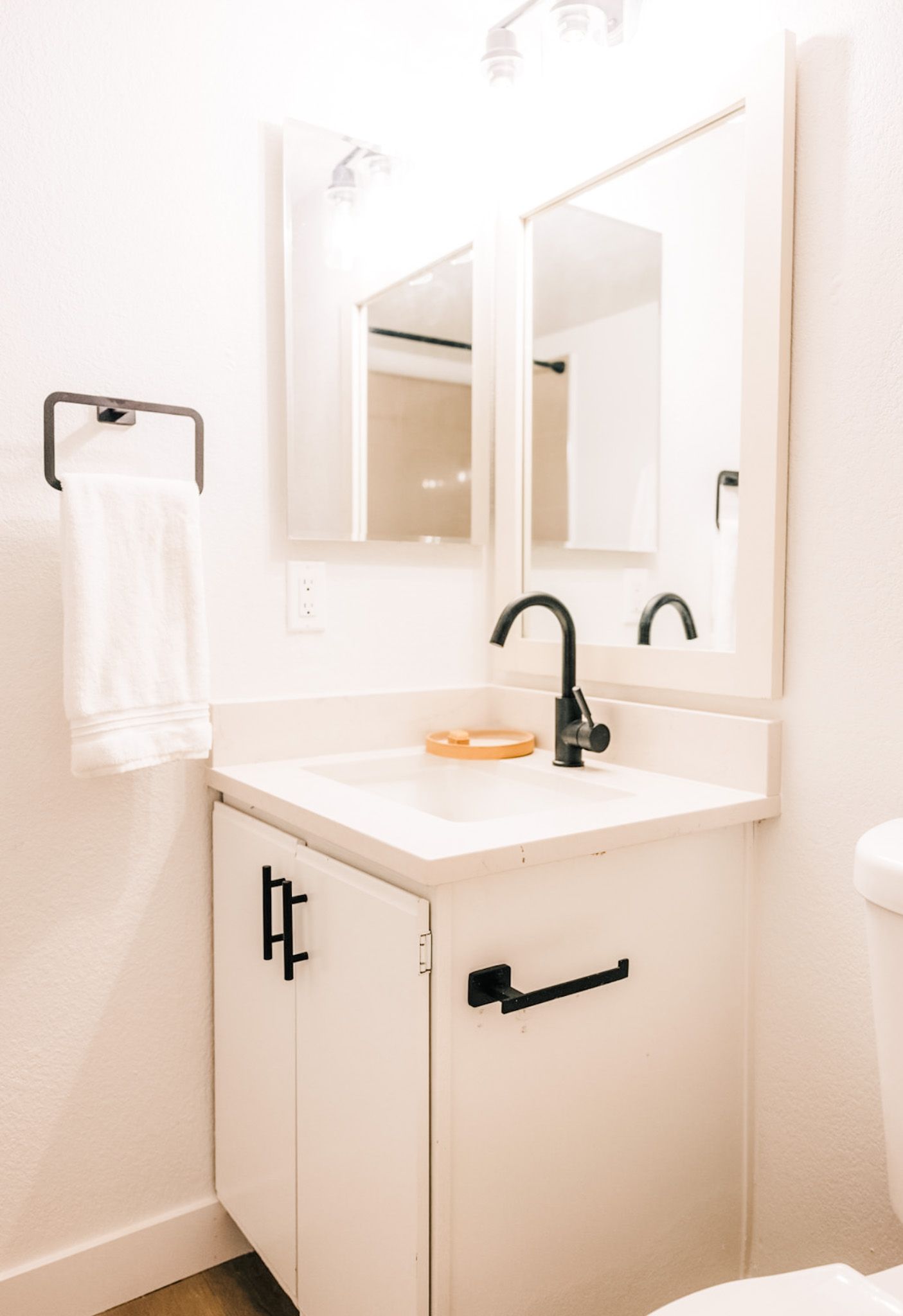 White bathroom with a sink, cabinet, and black fixtures. A towel hangs on a black rack.