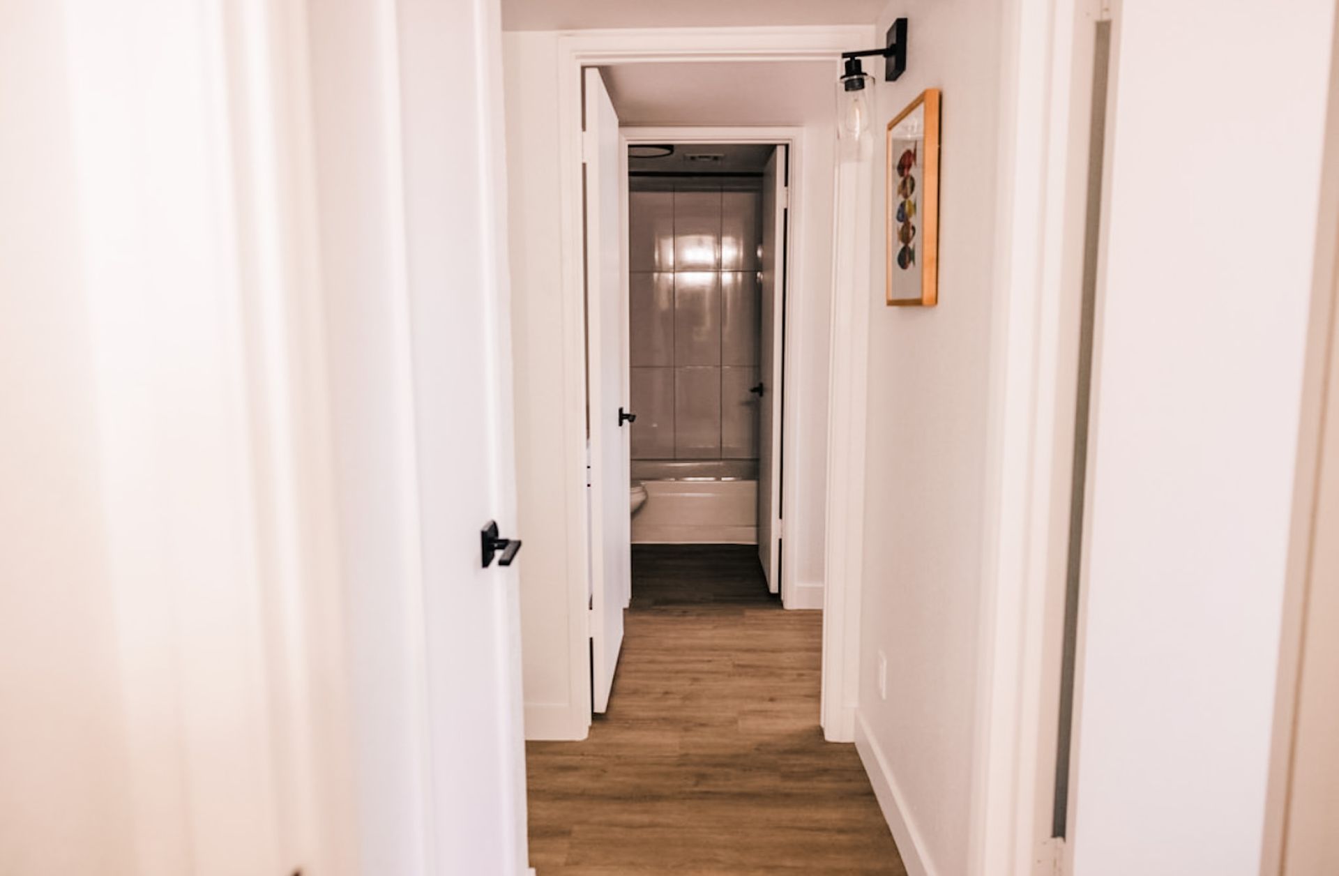 Hallway with white walls, wood floor, and open doors to a bathroom and a room.