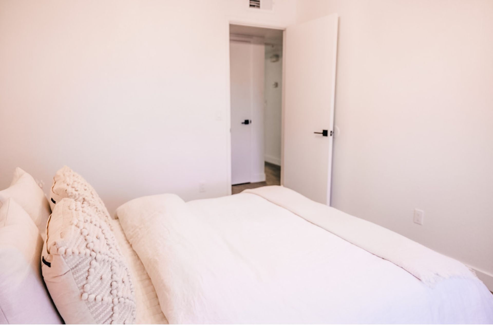 White bedroom with a bed, pillows, and an open door leading to another room.