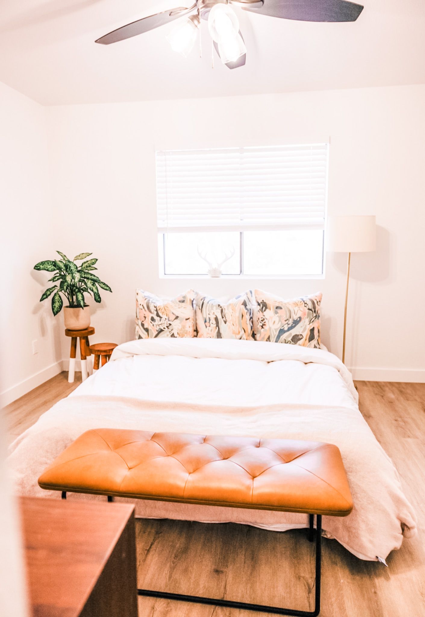 Bedroom with bed, bench, plant, and window with blinds. Neutral tones, wood flooring.