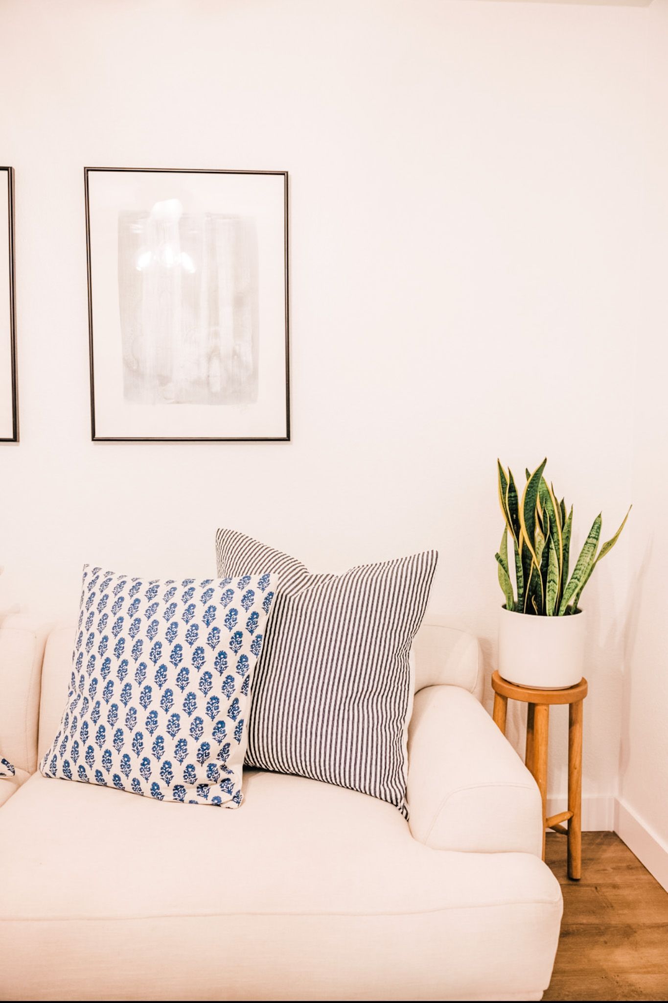 Cream-colored sofa with blue patterned pillows, framed art, and a snake plant on a wooden stand.