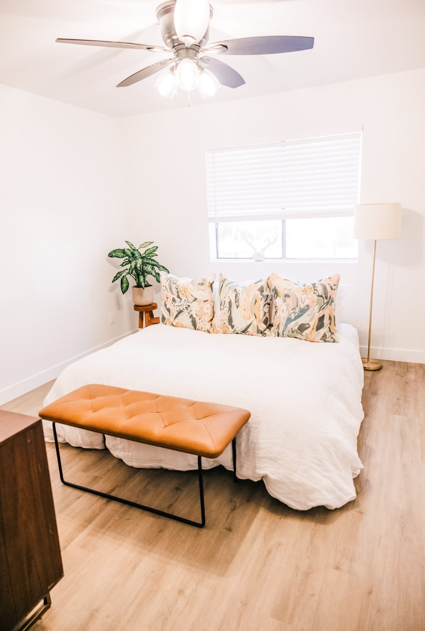 Bedroom with bed, bench, plant, and window with blinds; light wood floors, neutral tones.