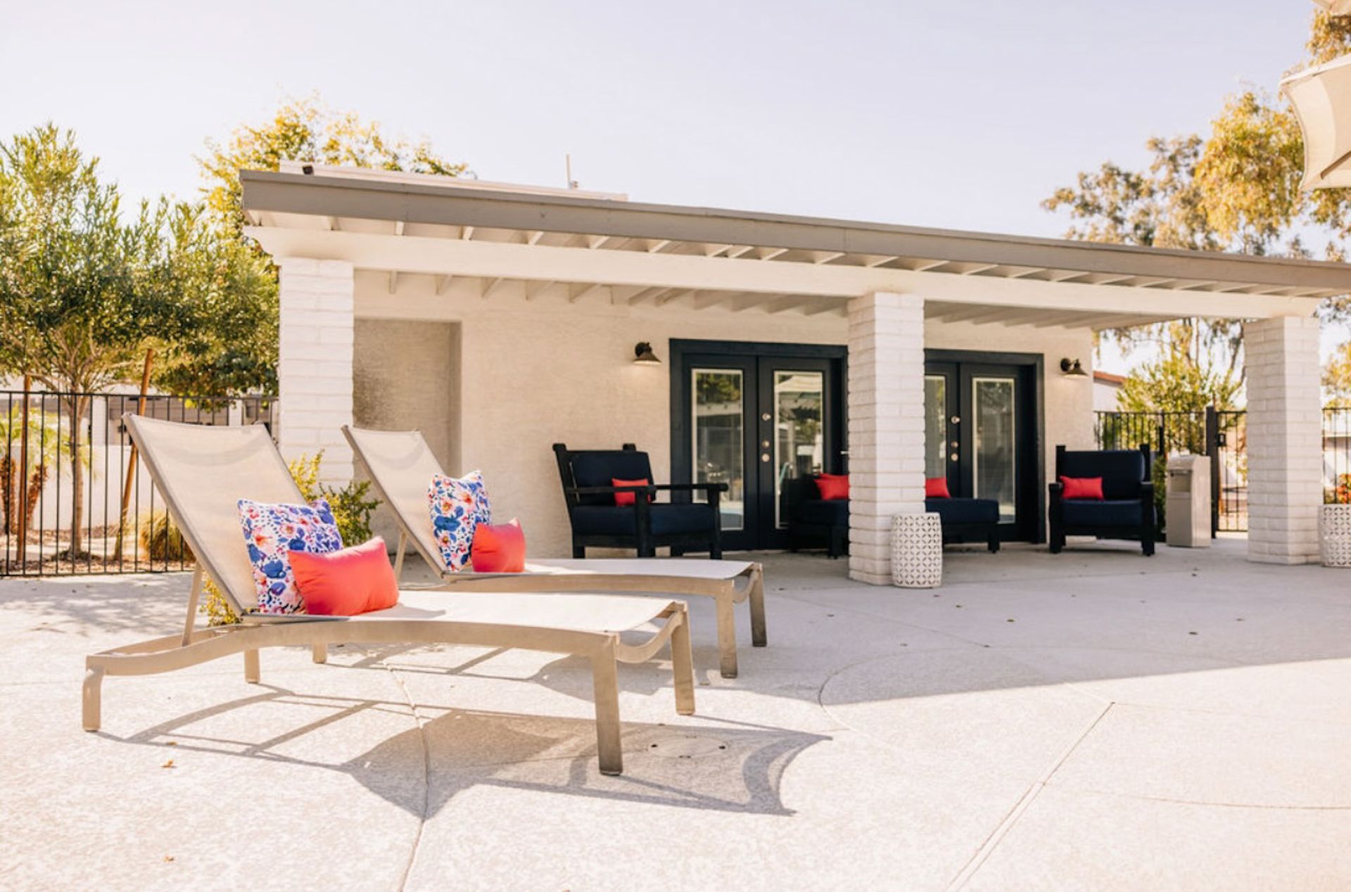 Two lounge chairs with red pillows face a poolside cabana with dark doors.