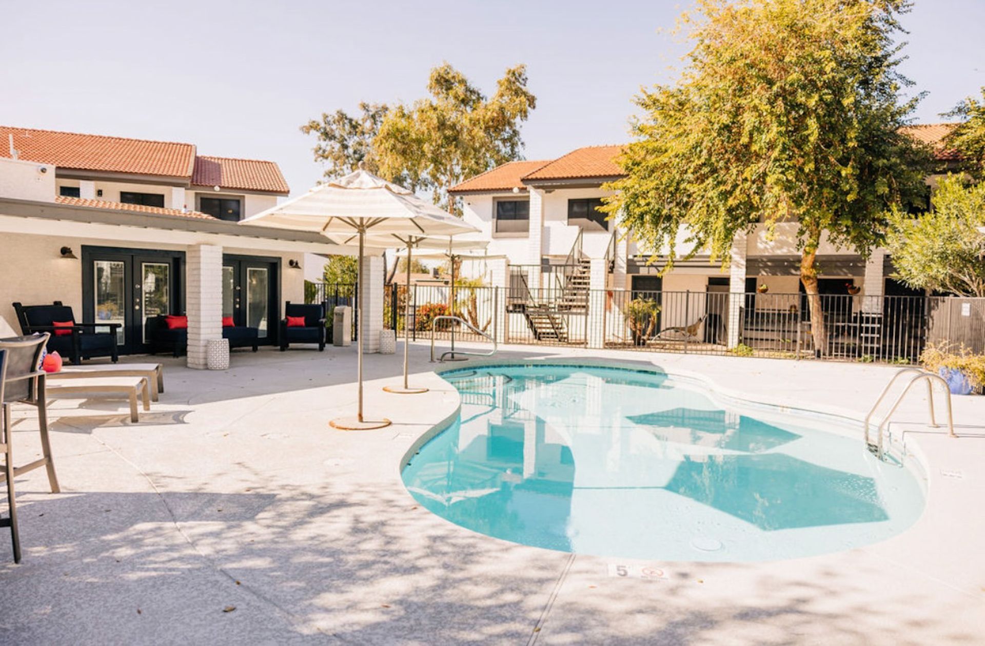 Pool area with white buildings, lounge furniture, and trees on a sunny day.