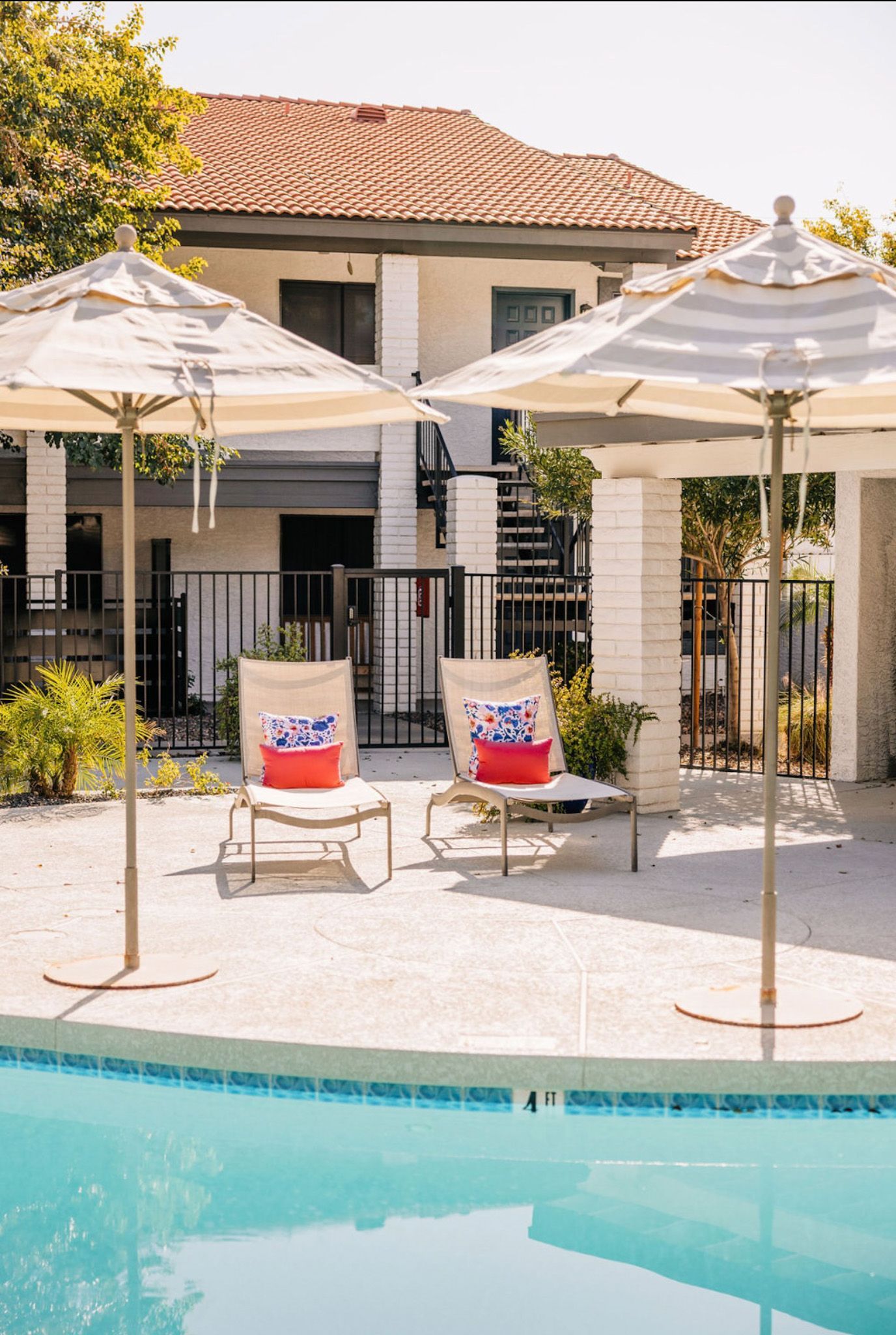 Two lounge chairs with red pillows sit near a pool under striped umbrellas; a building is in the background.