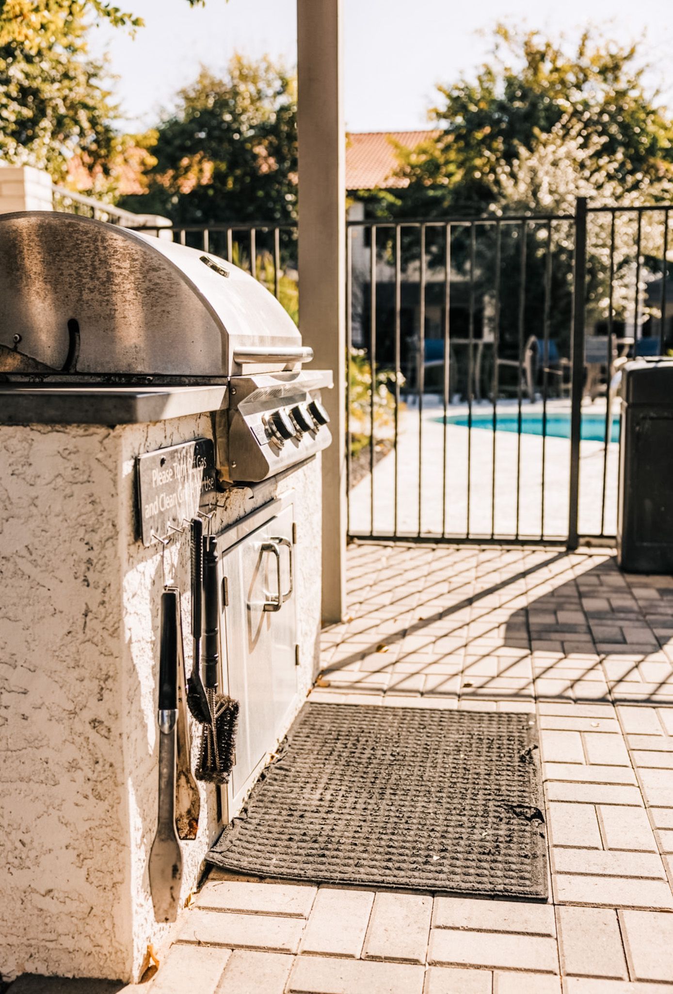 Outdoor grill on a brick patio, near a pool and gate, sunny.