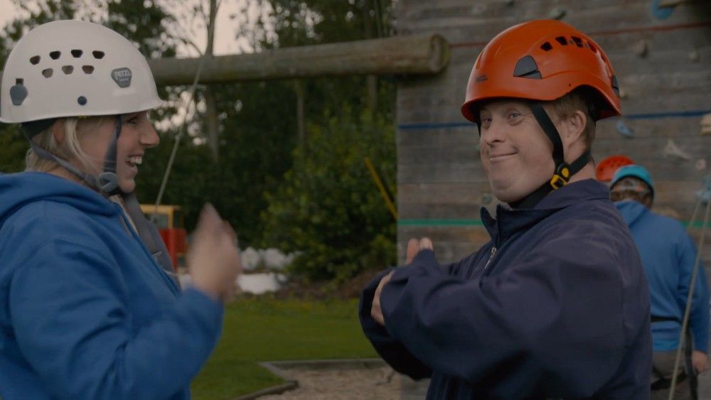 Two people in helmets and blue jackets smiling, clapping hands near a climbing wall.