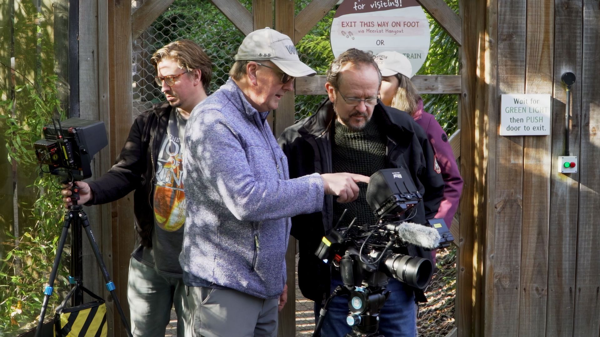 Filmmakers by a wooden gate, one points at a camera, others observe.