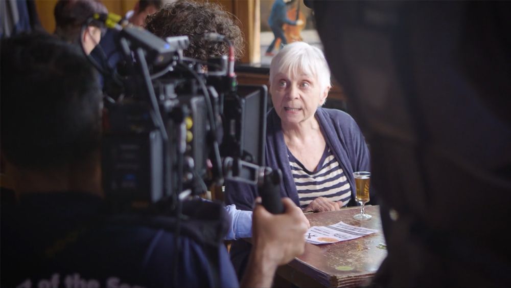 Woman in striped shirt being filmed, seated at a bar table. Camera equipment is in the foreground.