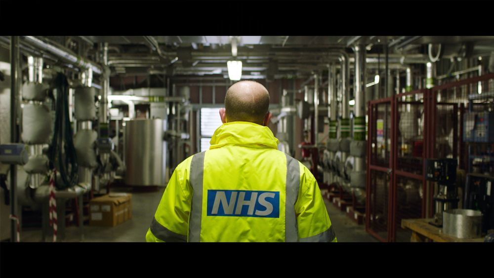 Man in high-vis NHS jacket walks through industrial facility with pipes and tanks.