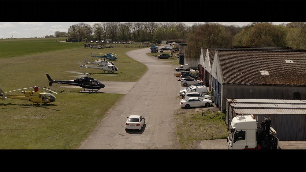 White car driving down a road at an airfield with helicopters and buildings.