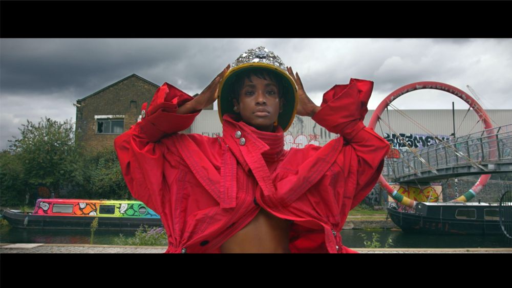 Person in red outfit, gold crown, hands on head. Canal setting, overcast sky, boats, graffiti.