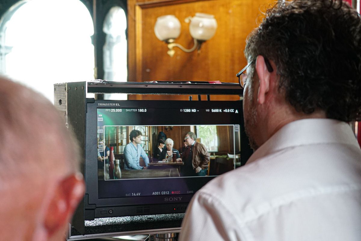 Person looking at a monitor displaying a scene with two men at a table. The setting appears to be indoors.