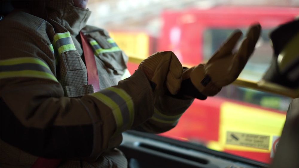 Firefighter in protective gear, inside a fire truck, gesturing with gloved hands.