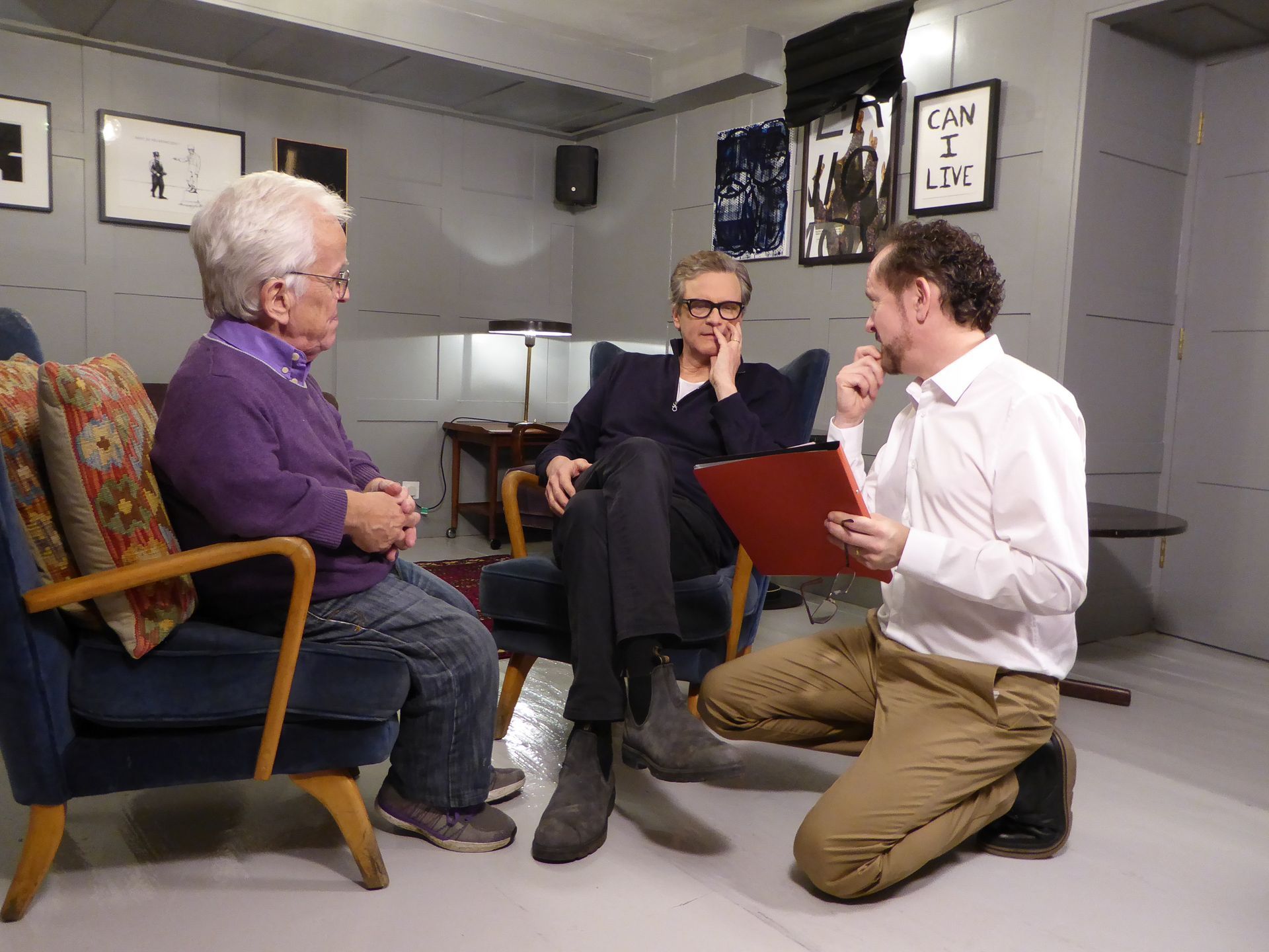 Three people in a room. One older person in a chair, two others seated and kneeling, possibly an interview or discussion.
