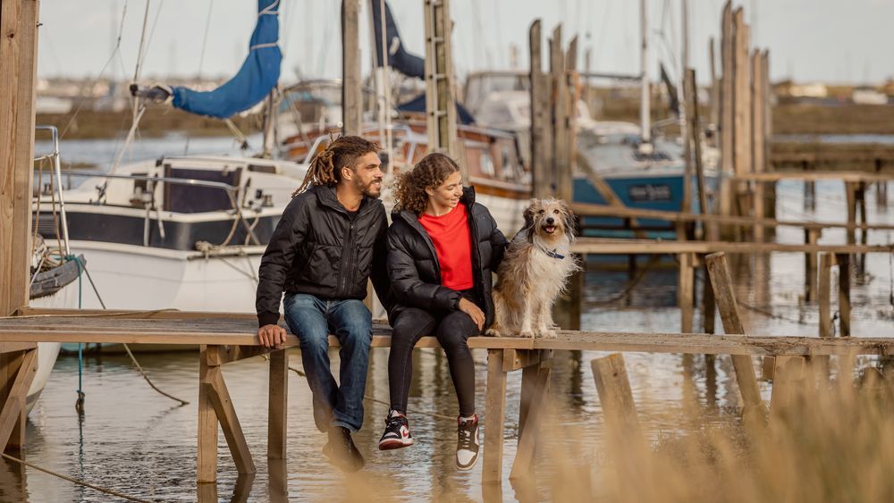 Couple and dog sit on a pier, overlooking boats. Man with dreadlocks and woman in jackets.