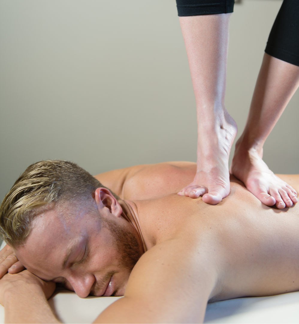 Man receiving back massage with therapist's feet; tan skin, neutral background.