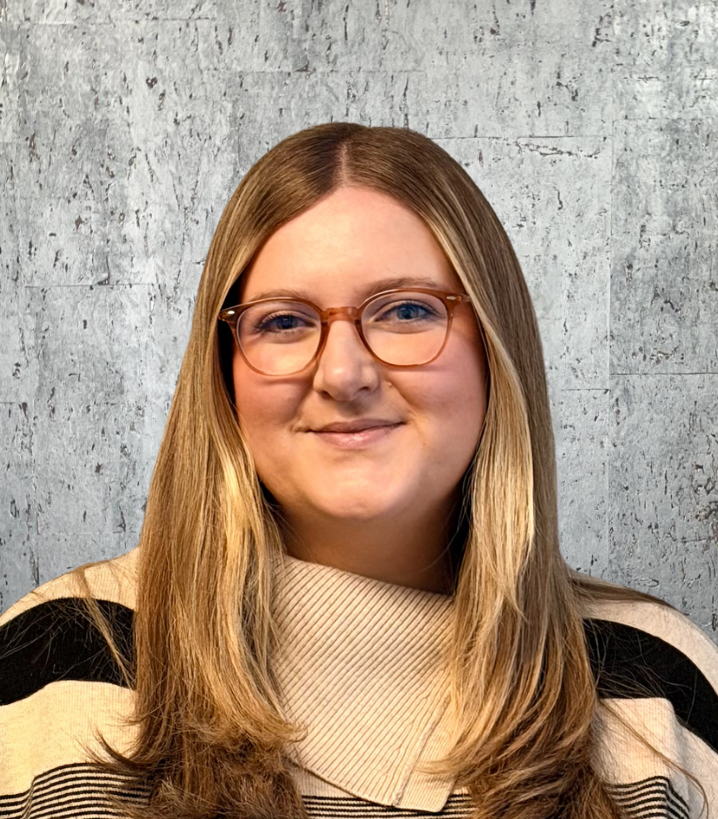 Woman with long blonde hair, glasses, wearing a light-colored sweater, standing in front of a gray wall.