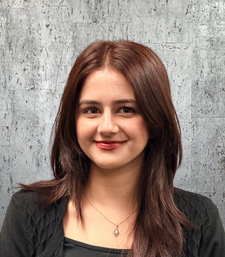 Woman with brown hair smiles, wearing a black top and pendant, against a textured gray wall.